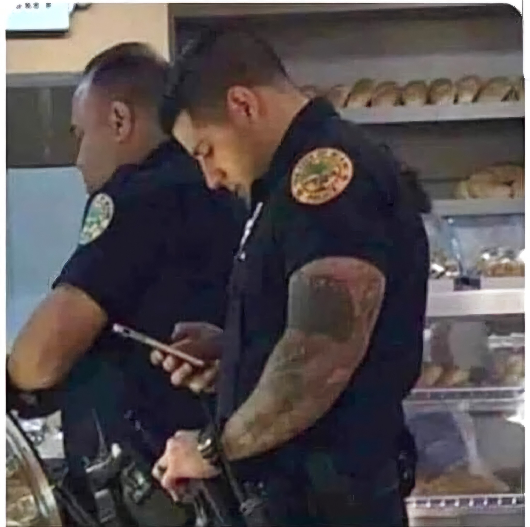 Dva policajti v uniforme pri pulte s pečivom, jeden z nich kontroluje svoj telefón.