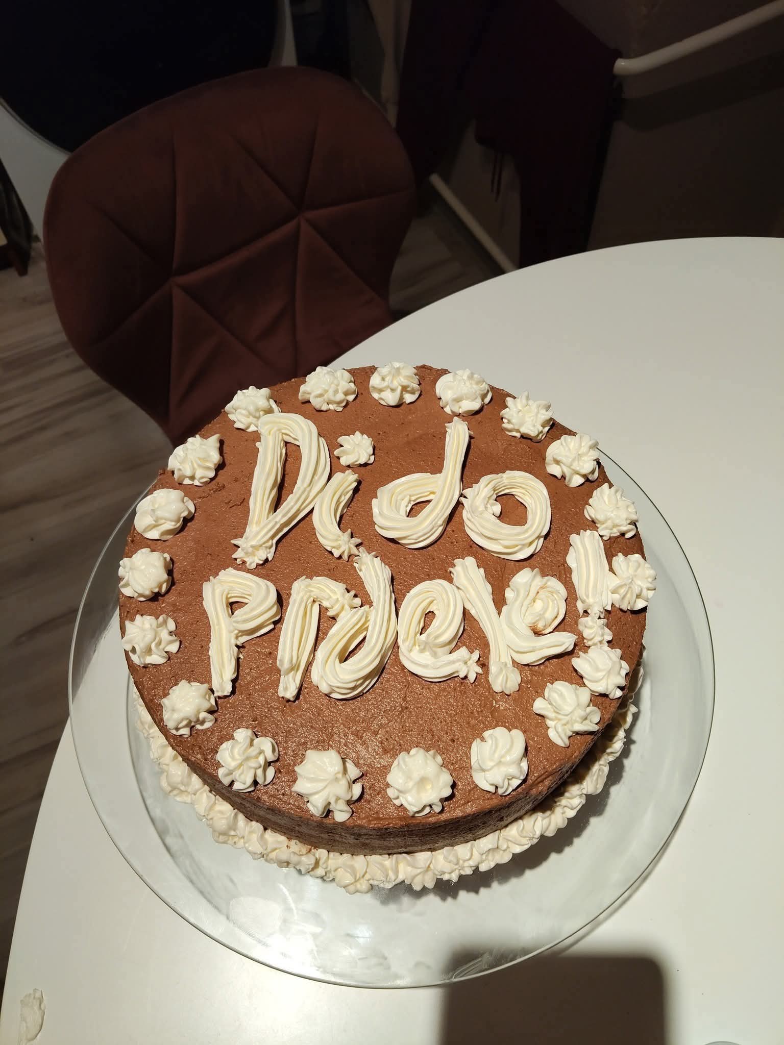 Čokoládová torta s nápisom Dedo prdeľ ozdobená krémom na bielom stole a pri hnedej stoličke.