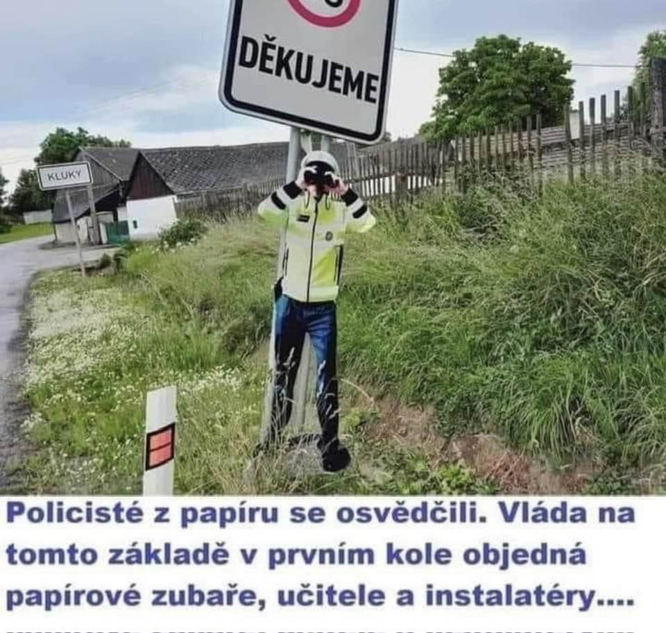 Papierový policajt s ďalekohľadom pri ceste a značení rýchlosti, plagát s nápisom DĚKUJEME, dedina v pozadí.