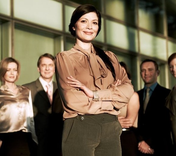 Skupina profesionálov stojí pred modernou budovou, žena vpredu s úsmevom a prekríženými rukami.