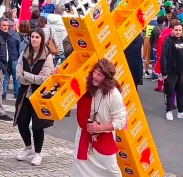 Muž v kostýme Krista nesie kríž z pivových dební počas karnevalu na rušnej ulici, okolo prechádza množstvo ľudí.