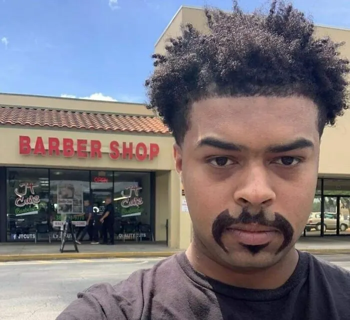 Muž s unikátnym fúzovým štýlom pred holičstvom, exteriér budovy.