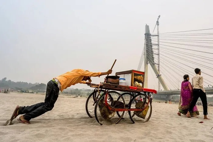 Muž tlačí vozík na piesku pri moste, zatiaľ čo pár kráča po pláži v pozadí.
