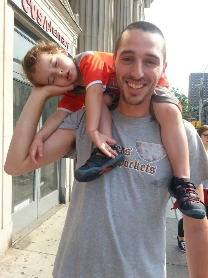 Otec s úsmevom nesie spiace dieťa na ramenách, prechádzka mestom pozdĺž ulice s obchodom.