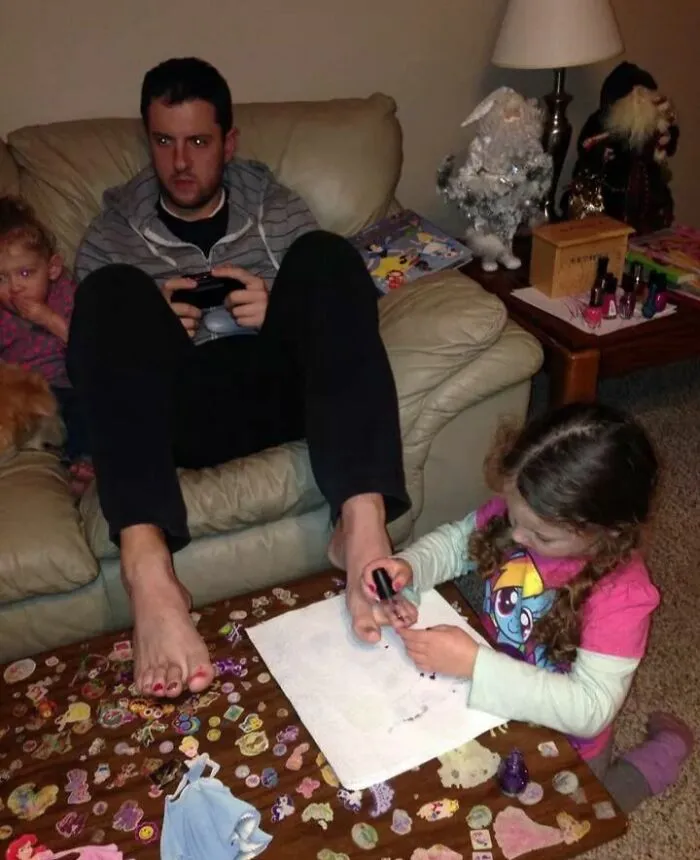 Dievčatko lakuje otcovi nechty na nohách, zatiaľ čo otec hrá videohru na konzole v obývačke.