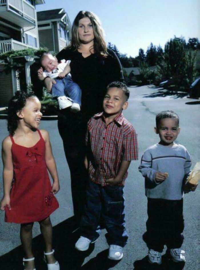 Žena so štyrmi deťmi stojí na ulici pred domami počas slnečného dňa, jedno dieťa je v náručí.