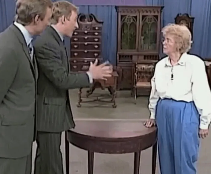 Ľudia diskutujú pri starožitnom stole na výstave antikvariátu, obklopení starožitným nábytkom na pozadí.