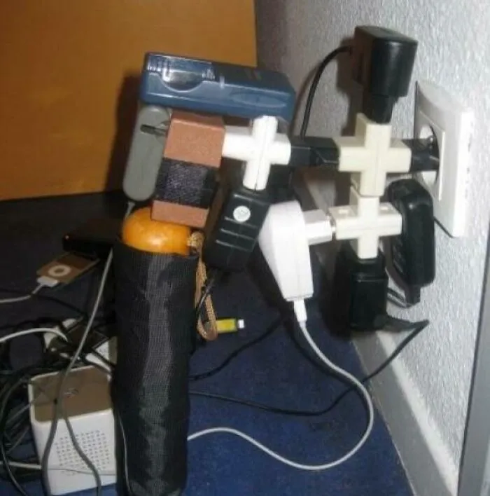 Nabíjačky a adaptéry preťažené v zásuvke s kreatívnym zapojením pomocou rozbočiek a podporené ovocím a dáždnikom.