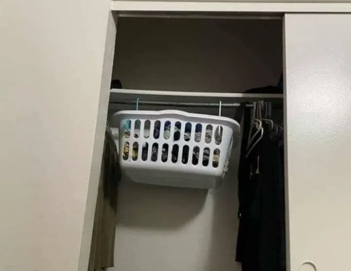 Biely košík plný oblečenia zavesený vo vstavanej skrini na kovovej šatníkovej tyči.