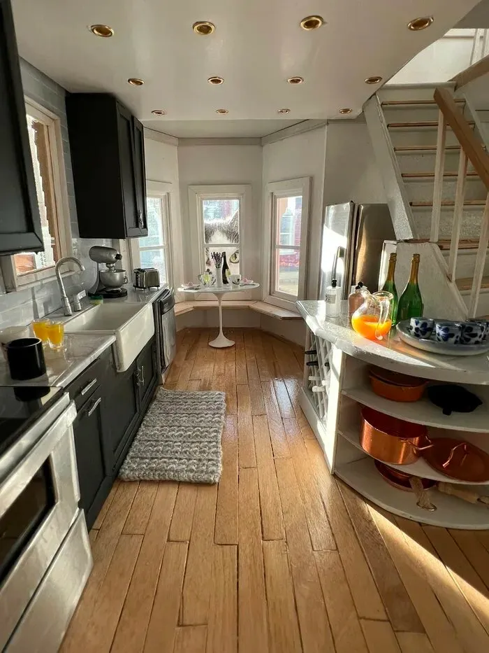 Moderná kuchyňa s drevenou podlahou, čiernymi skrinkami, bielymi stenami a okrúhlym stolom pri okne.