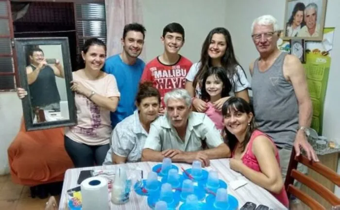 Skupina ľudí spolu trávi spoločný čas pri stole s pohármi, niektorí sa usmievajú a držia zrkadlo.