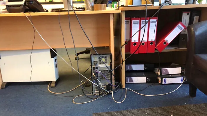 Stôl v kancelárii s počítačom, káble a šanóny v poličke, neorganizovaná elektroinštalácia.