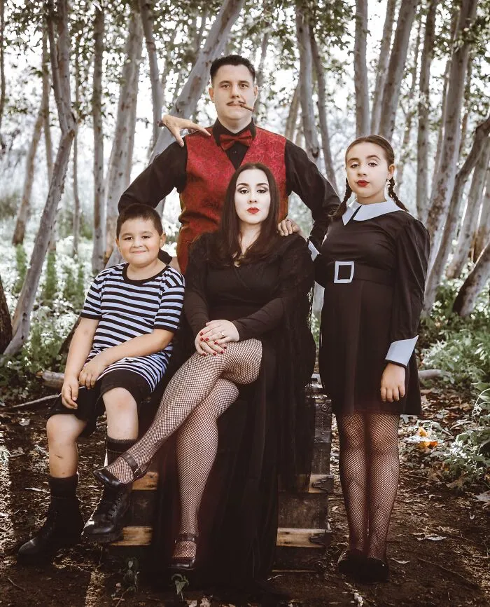 Rodina v kostýmoch stojí v lese, odetá v tmavých farbách, s dramatickým výrazom.