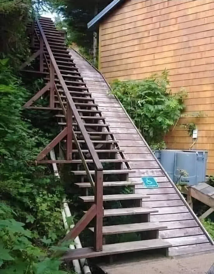 Rampy a schody v lese vedľa domu, s dopravnou značkou pre hendikepovaných používanou v nesprávnom kontexte.