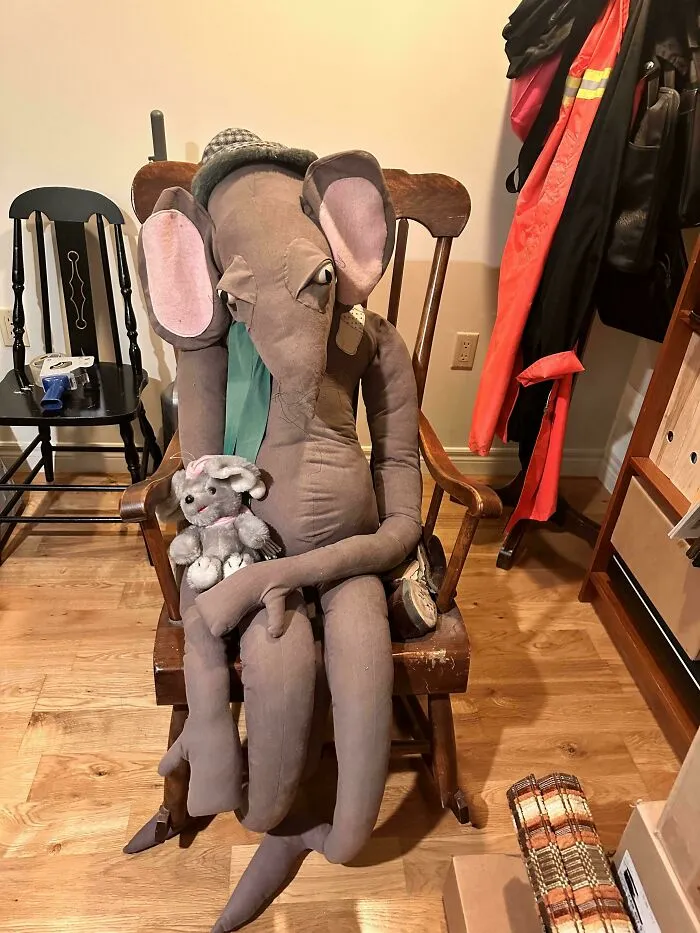 Plyšový slon s klobúkom sedí na drevenej stoličke, držiaci menšieho plyšového zajaca v interiéri.