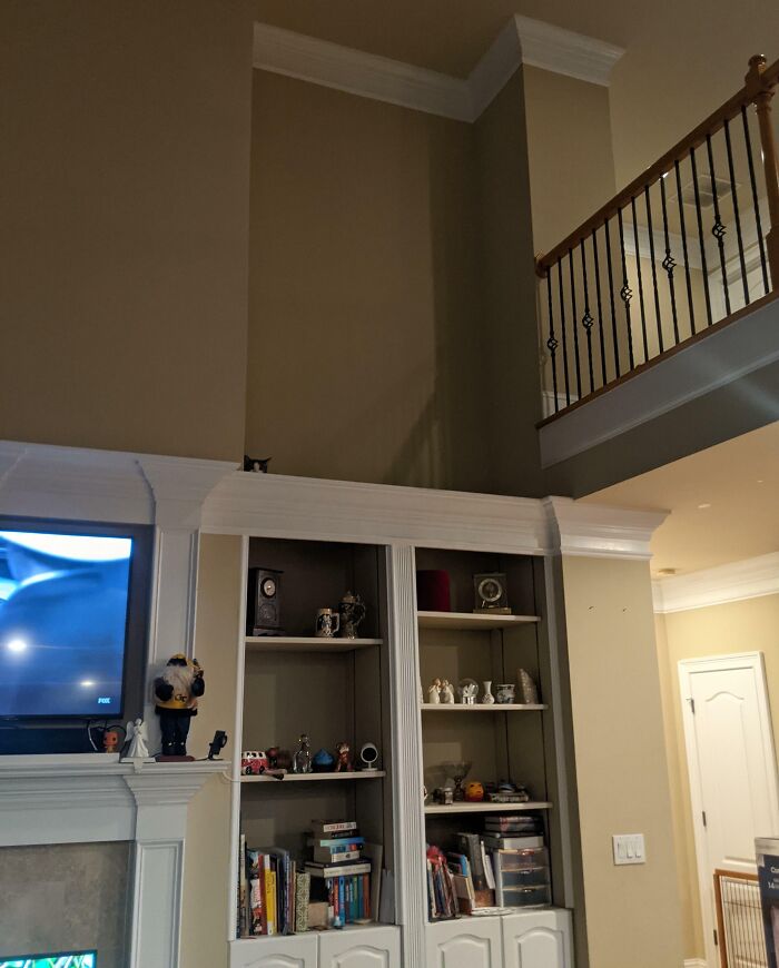 Obývacia izba s policou a skrytým mačacím pozorovateľom na vrchu, vedľa televízie.