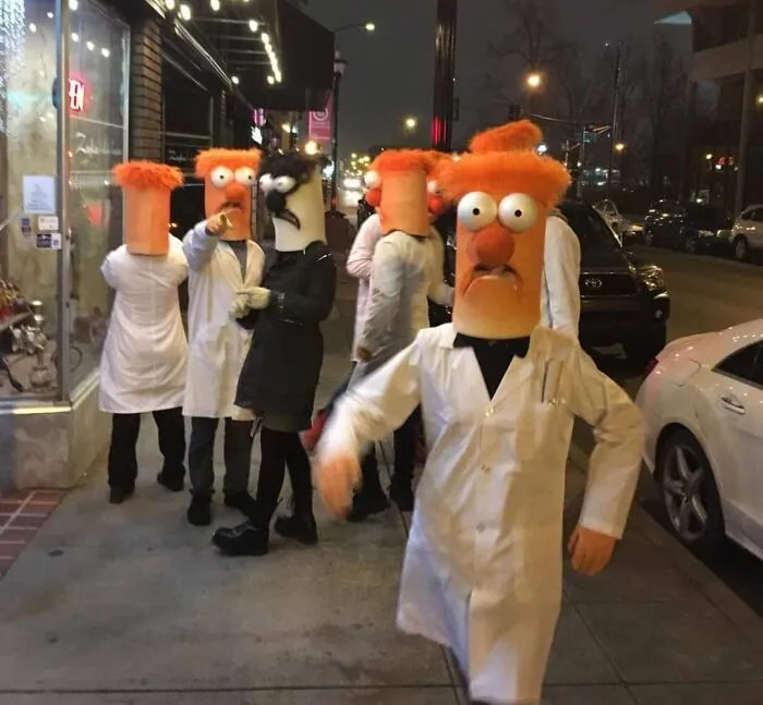 Skupina ľudí v kostýmoch postáv z Muppetov na večernej ulici v bielych plášťoch.