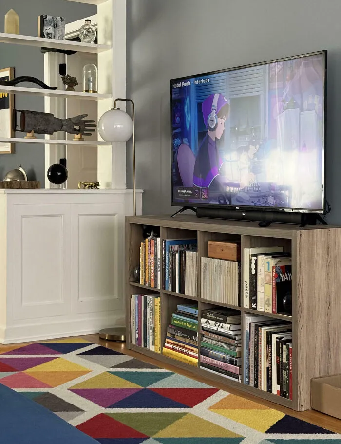 Obývačka s farebným kobercom, drevenou policou na knihy a TV s animovaným seriálom na obrazovke.