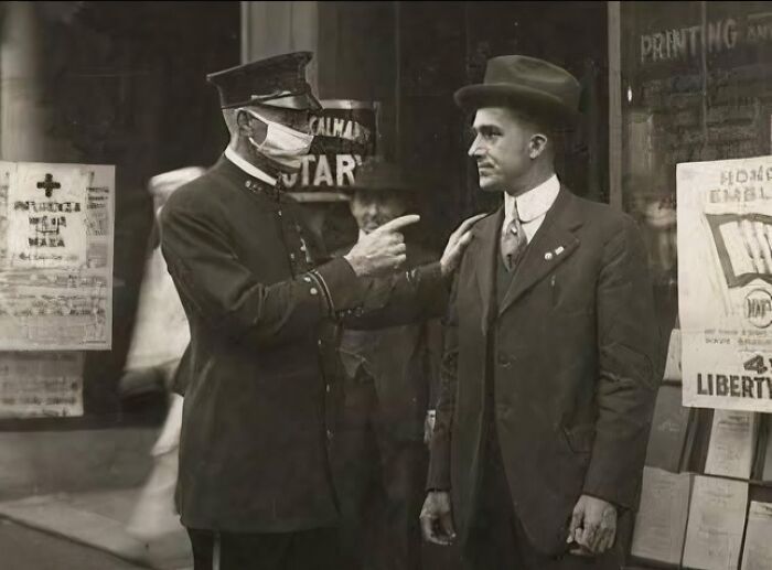 Policajt v rúšku a muž v klobúku diskutujú na ulici počas epidémie, historická fotografia.