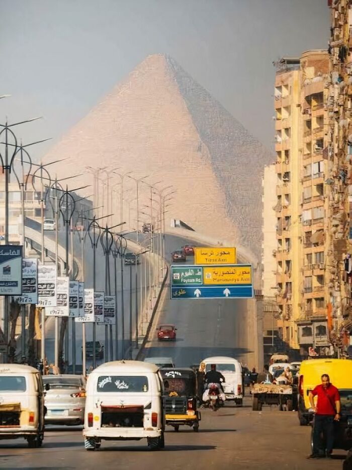 Mesto s premávkou a pyramídou v pozadí, mix starobylého a moderného prostredia pod jasnou oblohou.