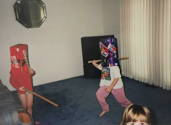 Deti s krabicami na hlavách a palicami v obývačke, hravá scéna plná fantázie a smiechu.