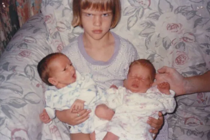 Dievčatko drží dve bábätká a tvári sa nespokojne, sedí na vzorovanej pohovke.