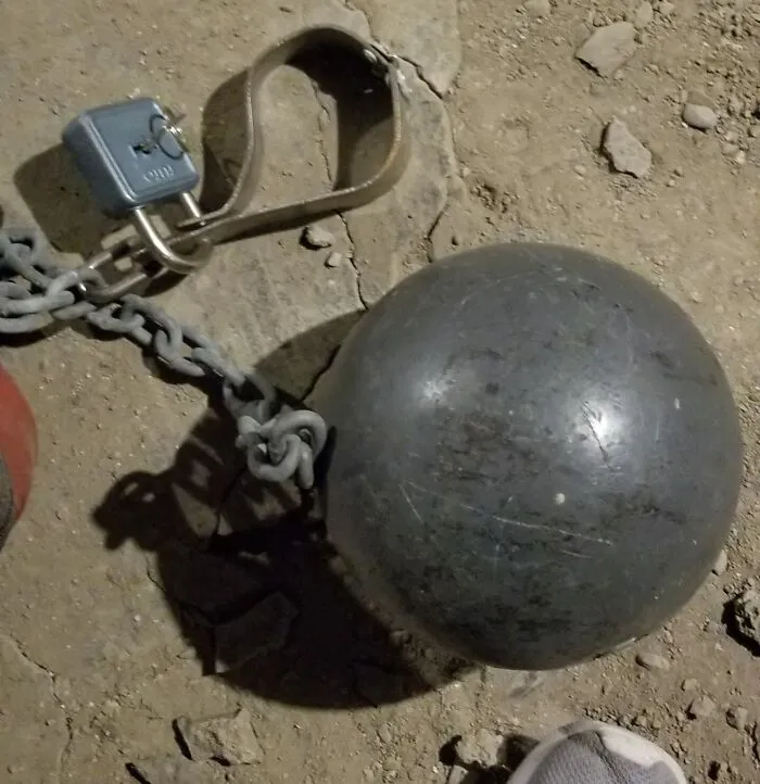 Železná guľa na reťazi s visacím zámkom na prašnej zemi, symbol väzenia a obmedzenia pohybu.