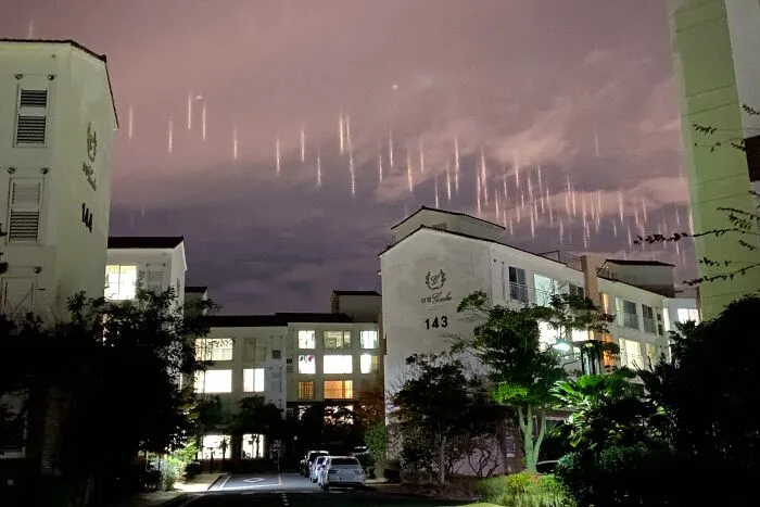 Nočná obloha nad osvetlenými budovami s padajúcimi svetelnými pruhmi v pozadí, pokojný mestský večer.