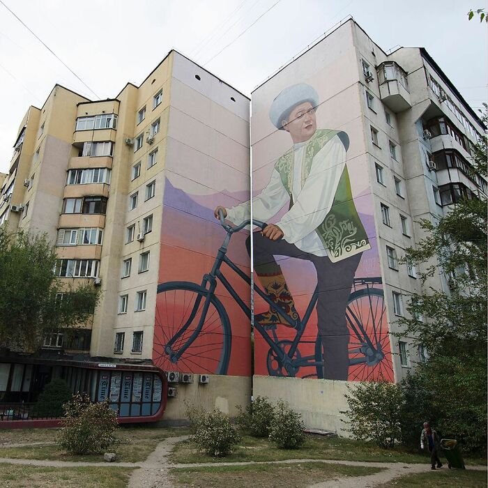 Veľký nástenný mural muža na bicykli v tradičnom ľudovom kroji na panelovom dome.