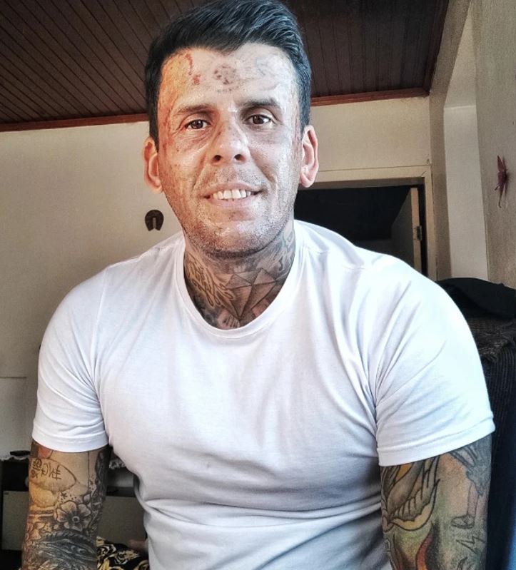 Osoba s tetovaniami oblečená v bielom tričku sedí v miestnosti a usmieva sa na kameru.