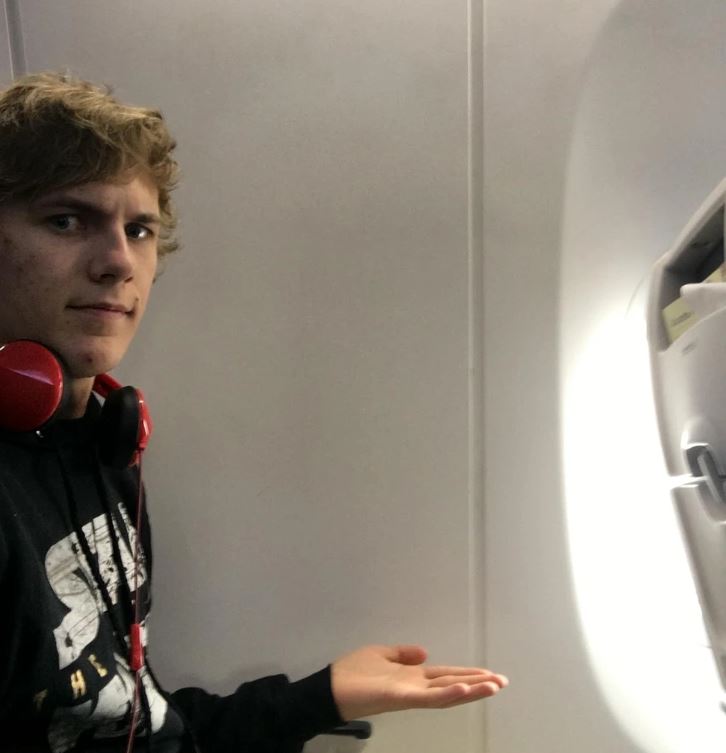 Osoba ukazujúca na stenu v interiéri lietadla s červenými slúchadlami okolo krku.