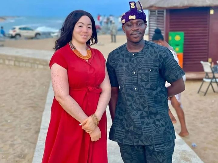 Dvojica v tradičnom oblečení stojí na pláži za slnečného dňa s výhľadom na oceán.