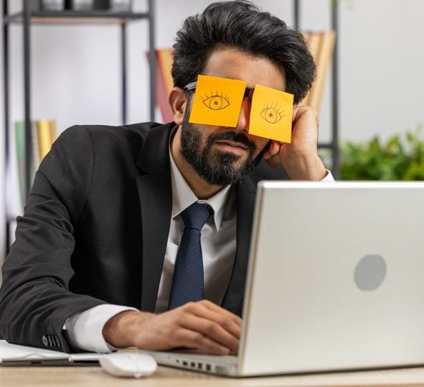 Muž znudene sedí pri notebooku s nalepenými papierikmi na očiach, ktoré ho imitujú bdelosť.