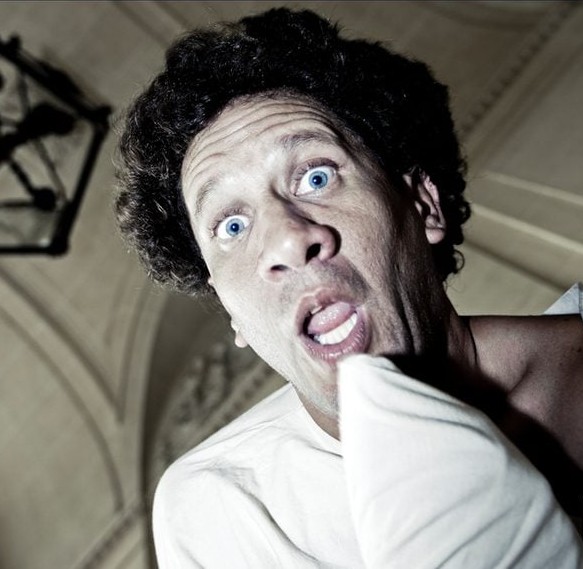 Muž s vystrašeným výrazom a modrými očami drží vankúš, interiér s vysokým stropom v pozadí.