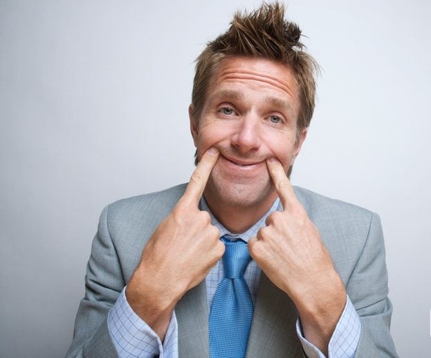 Muž v obleku s modrou kravatou si prstami vytvára úsmev na tvári, stojí proti svetlému pozadiu.