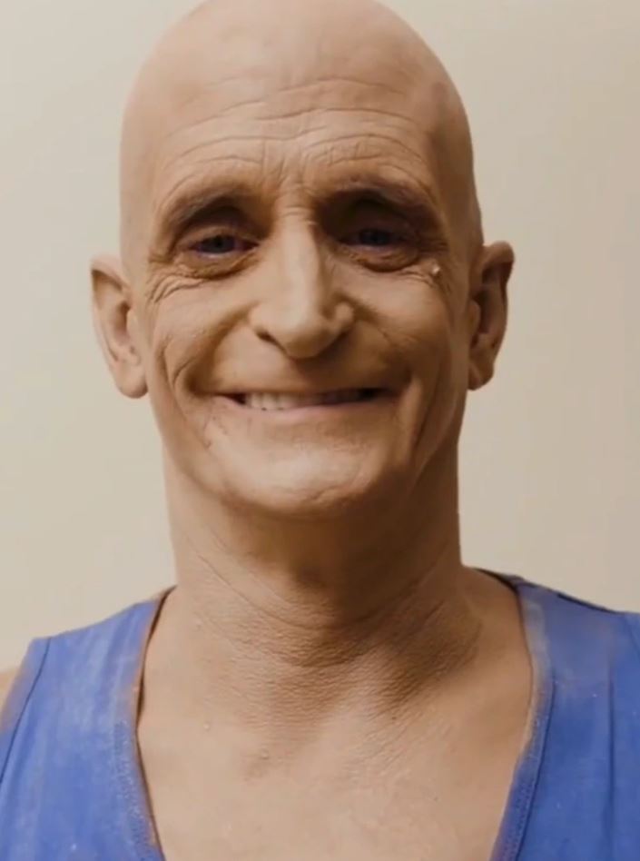 Usmievajúci sa muž s plešatou hlavou v modrom tričku pred béžovým pozadím.