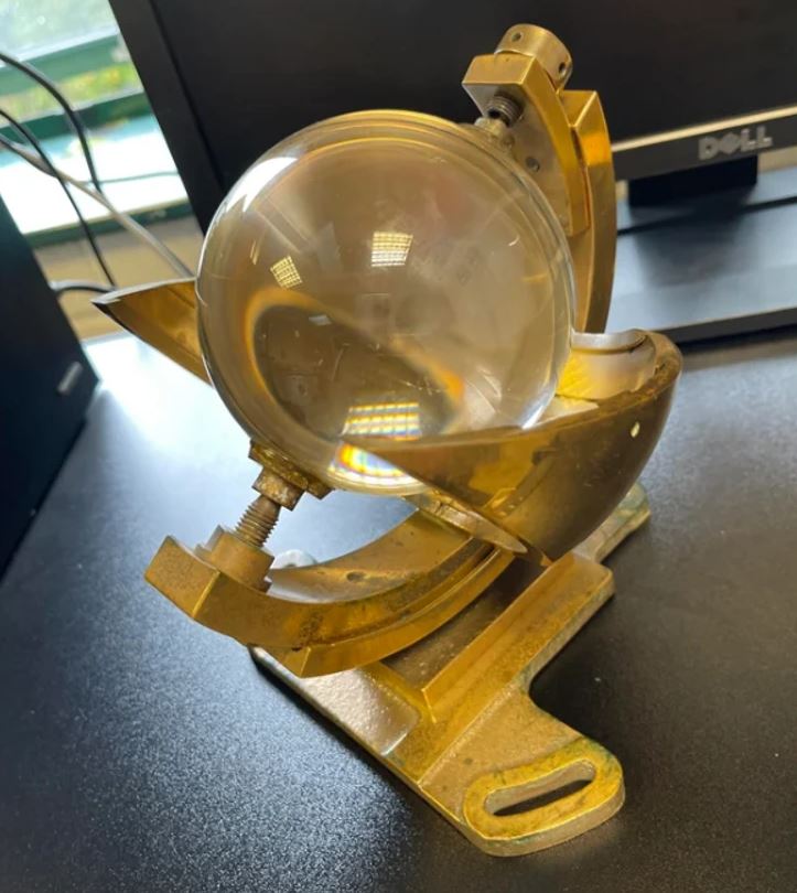 Starodávny zariadenie na meranie slnečného žiarenia na tmavom stole, okolo sú kancelárske počítače.