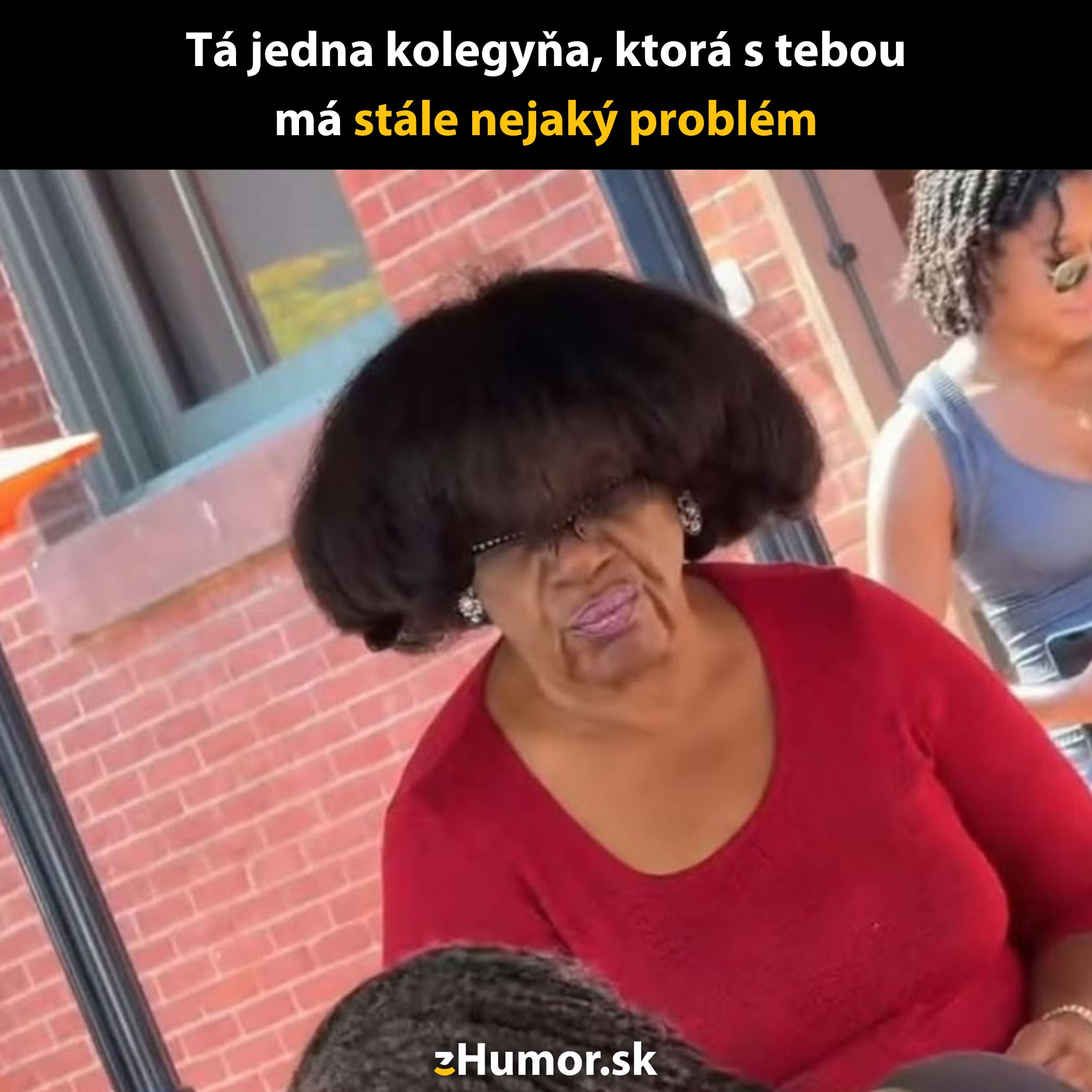 Neznáma žena v červenej blúzke s výrazným účesom stojí vonku pri tehlovej stene.