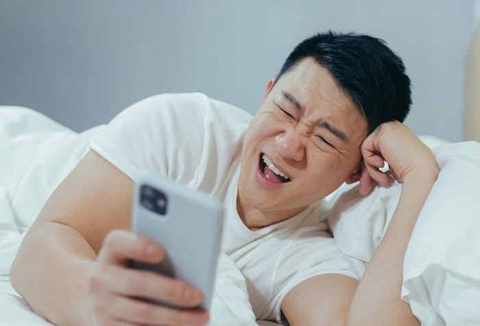 Muž ležiaci na posteli sa smeje, zatiaľ čo sa pozerá na telefón.