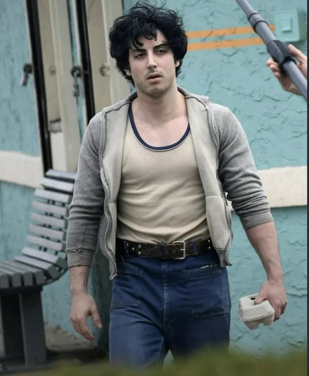 Muž v šedej mikine a džínsoch kráča po ulici, nesúc žltú škatuľu, na pozadí je modrá stena a lavička.