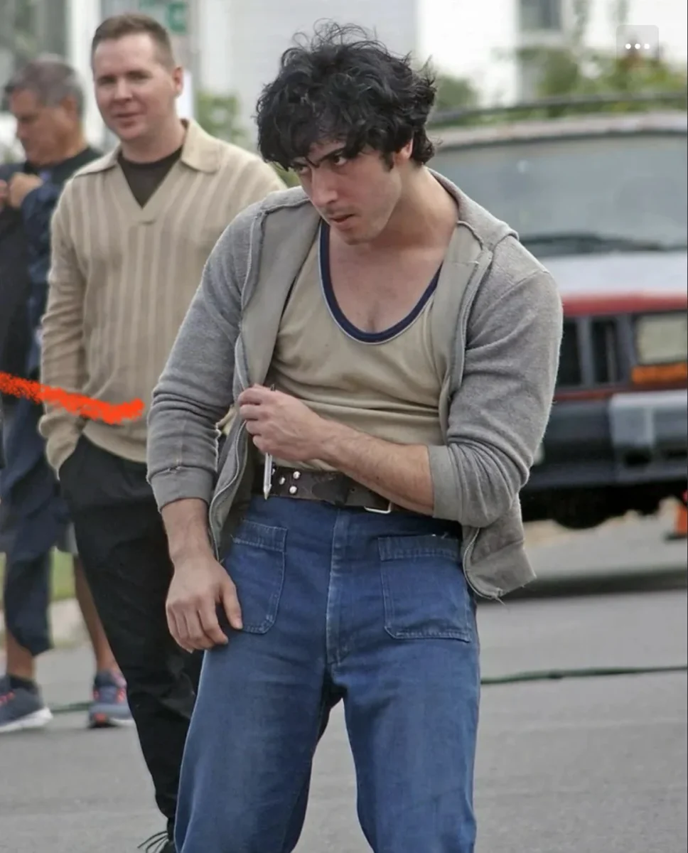 Muž vo svetri a džínsoch so zamysleným pohľadom stojí na ulici, v pozadí ďalší ľudia a auto.