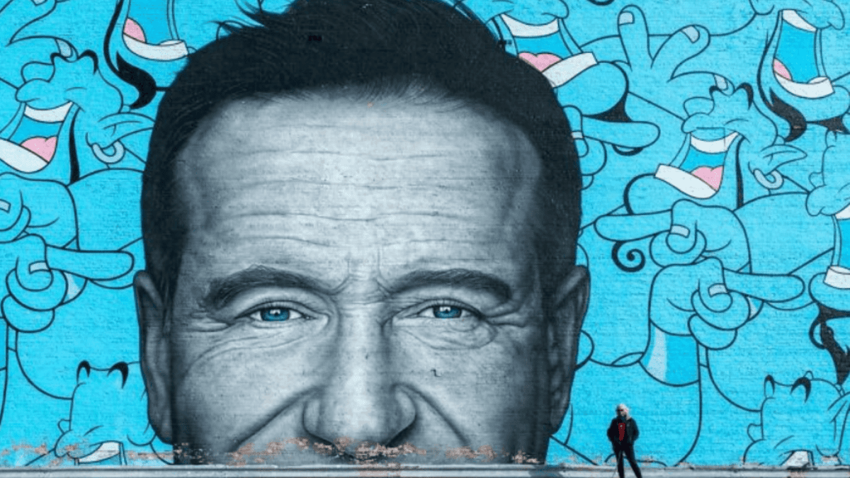 Nástenná maľba s tvárou muža a modrým pozadím plným veselých postavičiek, vedľa stojí človek v červenej bunde.