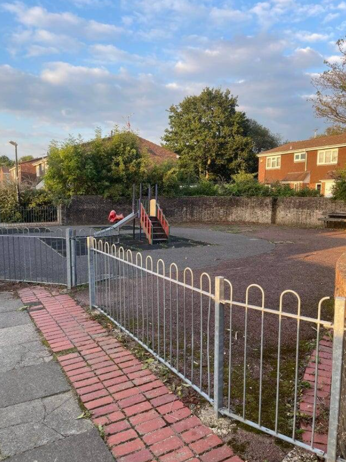 Detské ihrisko s preliezačkou, oplotenie, strom, obytné domy, modrá obloha s oblakmi v pozadí.