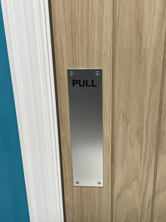 Dverový ťahací panel označený PULL na drevených dverách s kovovou doskou.