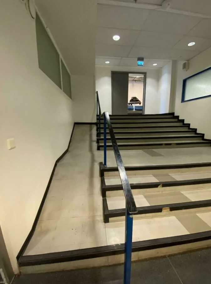 Schodisko s rampou v interiéri budovy, osvetlené stropnými svetlami, s cestou vedúcou k zatvoreným dverám.