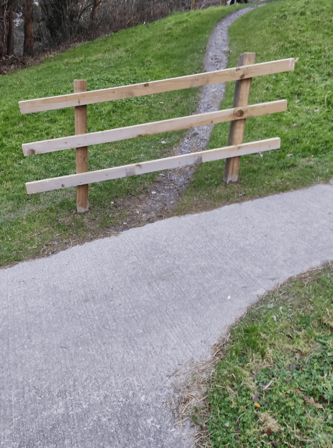 Drevený plot blokujúci cestu vedúcu z betónového chodníka na trávnatú plochu s úzkym blatistým chodníkom.