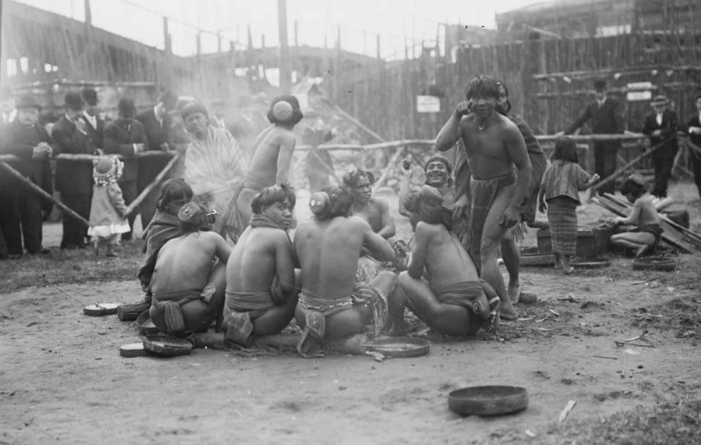 Skupina ľudí v tradičnom oblečení sedí, varí nad ohňom, zatiaľ čo sa okolo zbiehajú zvedaví diváci.