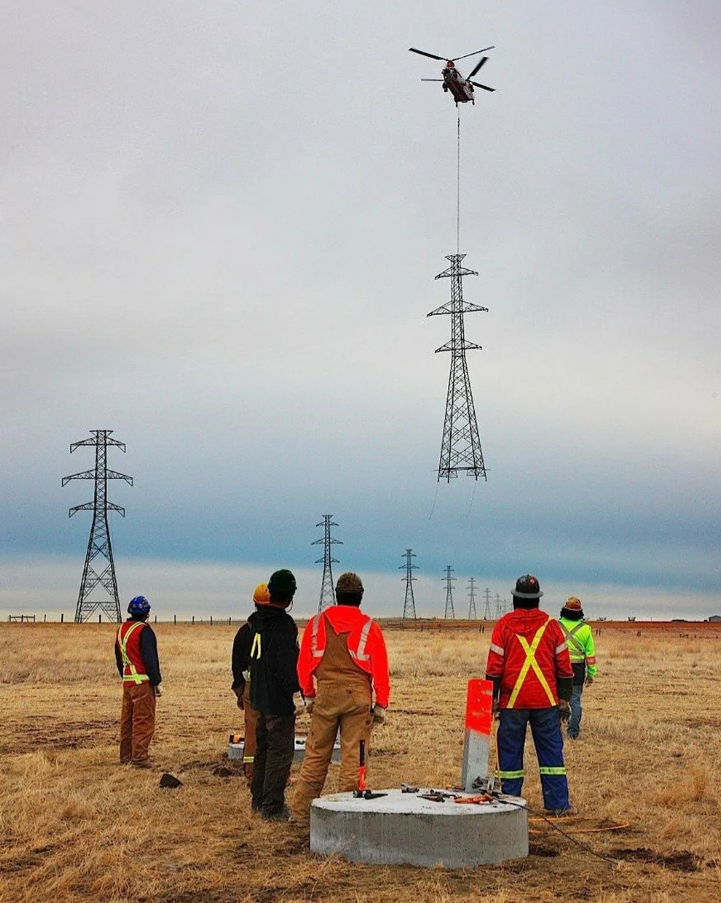 Helikoptéra inštaluje stĺp elektrického vedenia, pracovníci ho sledujú na poli.