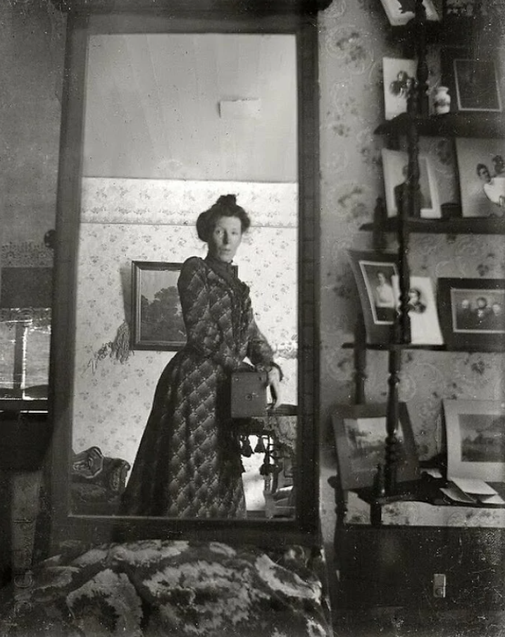 Žena v historických šatách sa fotografuje v zrkadle, vintage štýl, stará izba s dekoráciami.