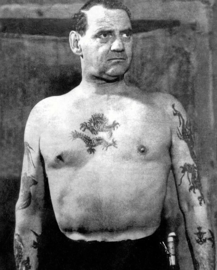 Odhodlaný muž s tetovaniami na hrudi a rukách stojí so sústredeným výrazom.